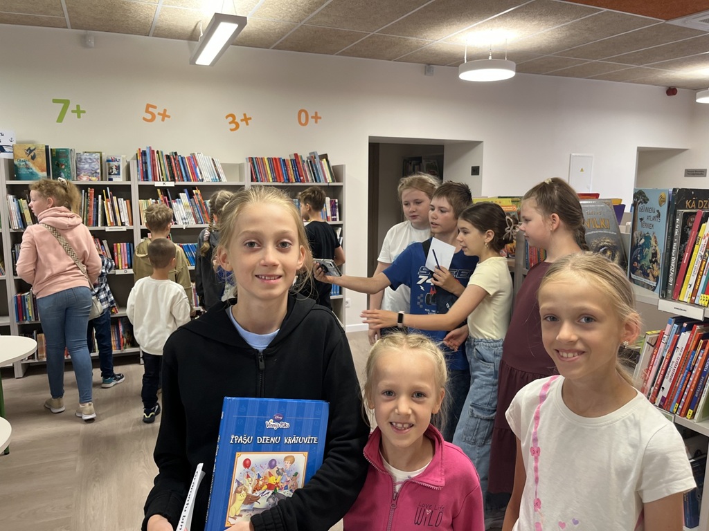 Trīs meitenes smaida un pozē priekšplānā bibliotēkā, kamēr viena no tām tur grāmatu "Īpašu dienu krātuvīte", un fonā pie plauktiem redzami citi bērni.