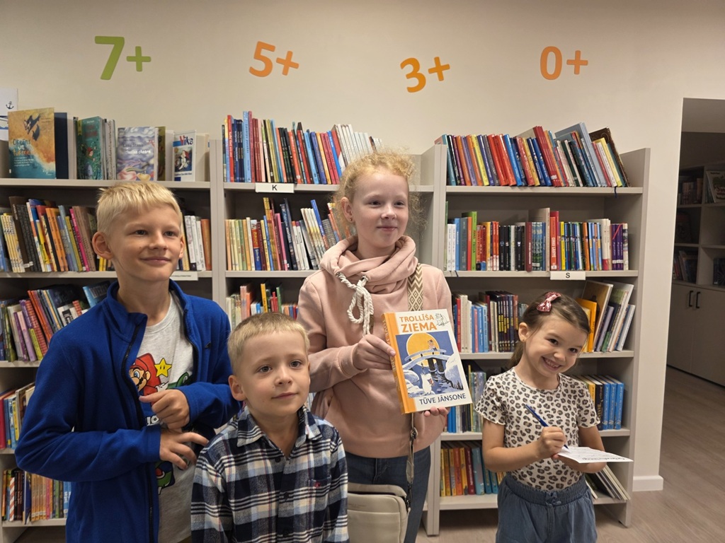 Četri bērni pozējot kamerai stāv pretī grāmatu plauktiem, kur virs tiem redzami vecuma apzīmējumi, un viena no meitenēm tur rokās grāmatu "Trollīša žiema".