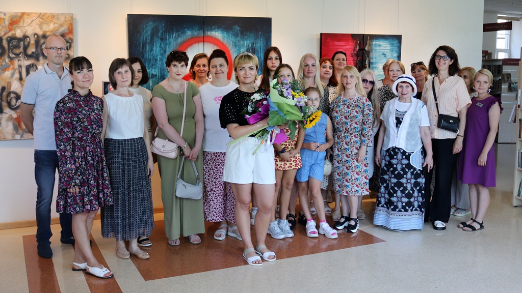 Liela cilvēku grupa kopīgi pozē piemiņas fotogrāfijai mākslas izstādē; centrā sieviete tur krāsainu ziedu pušķi, apkārt stāv gan sievietes, gan vīrieši un bērni.