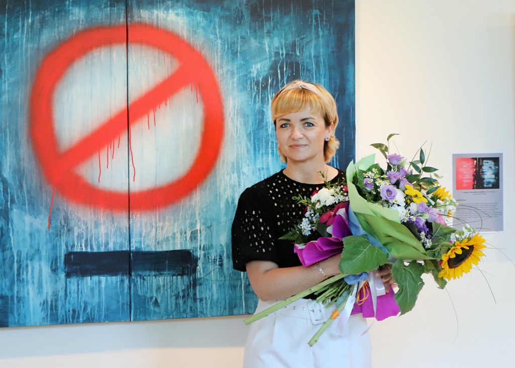Blonda sieviete melnā džemperī ar īsām piedurknēm un baltos šortos stāv izstādē un tur lielus krāsainu ziedu pušķus, fonā redzama glezna ar sarkanu aizlieguma zīmi uz zila fona.