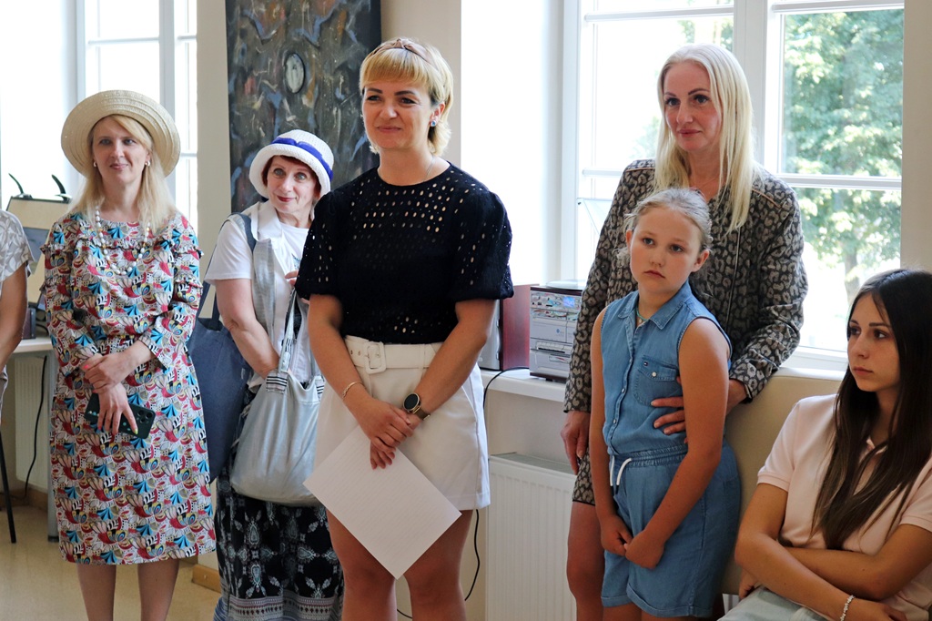 Grupa cilvēku stāv telpā, priekšplānā redzama blonda sieviete melnā džemperī ar dokumentiem rokās, blakus stāv sieviete ar cepuri, meitene džinsa kostīmā un citi apmeklētāji.
