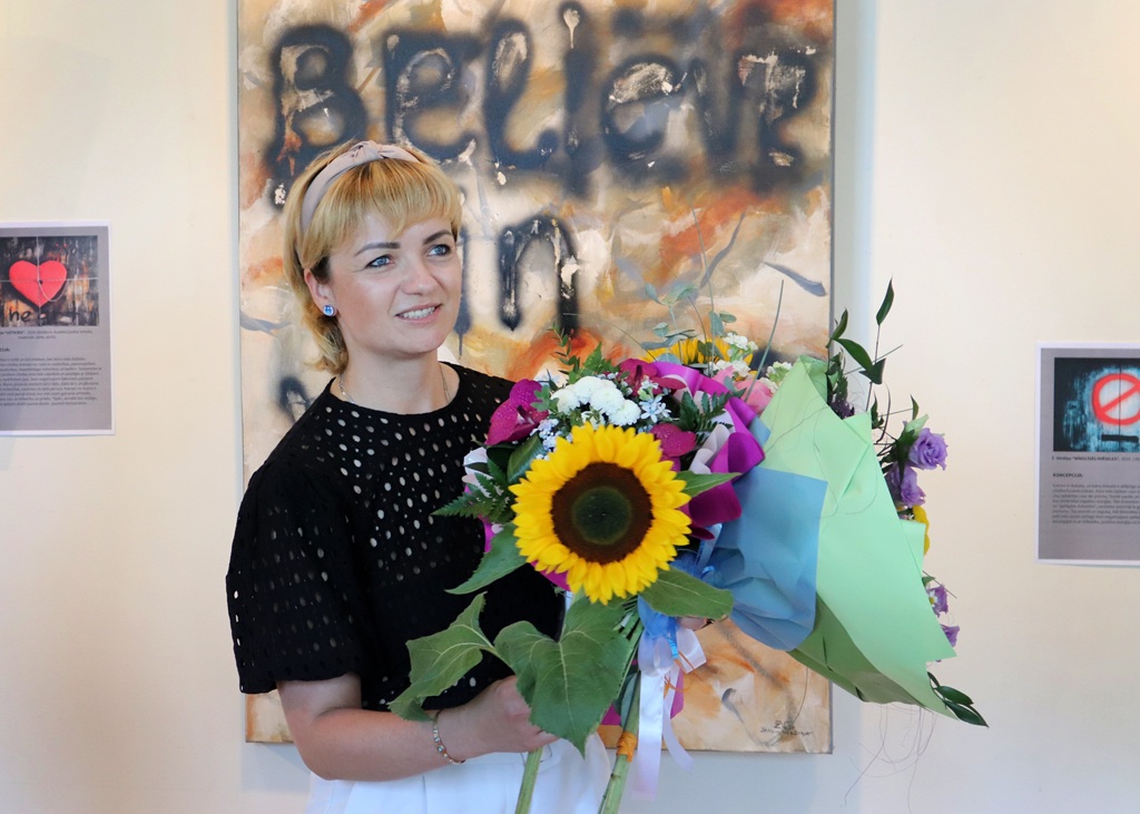 Blonda sieviete melnā džemperī smaidot tur lielu, krāsainu ziedu pušķi ar saulespuķi priekšplānā; fonā redzama glezna ar uzrakstu angļu valodā un gleznu apraksti piestiprināti pie sienas.