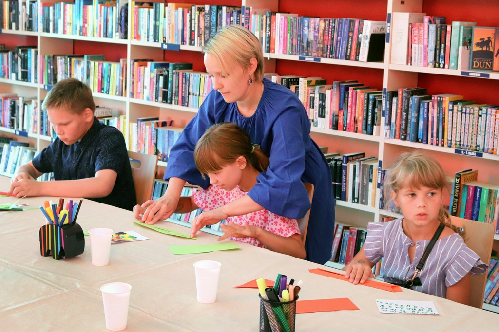 Bibliotēkas darbiniece palīdz meitenei veidot rokdarbus bērnu radošajā darbnīcā