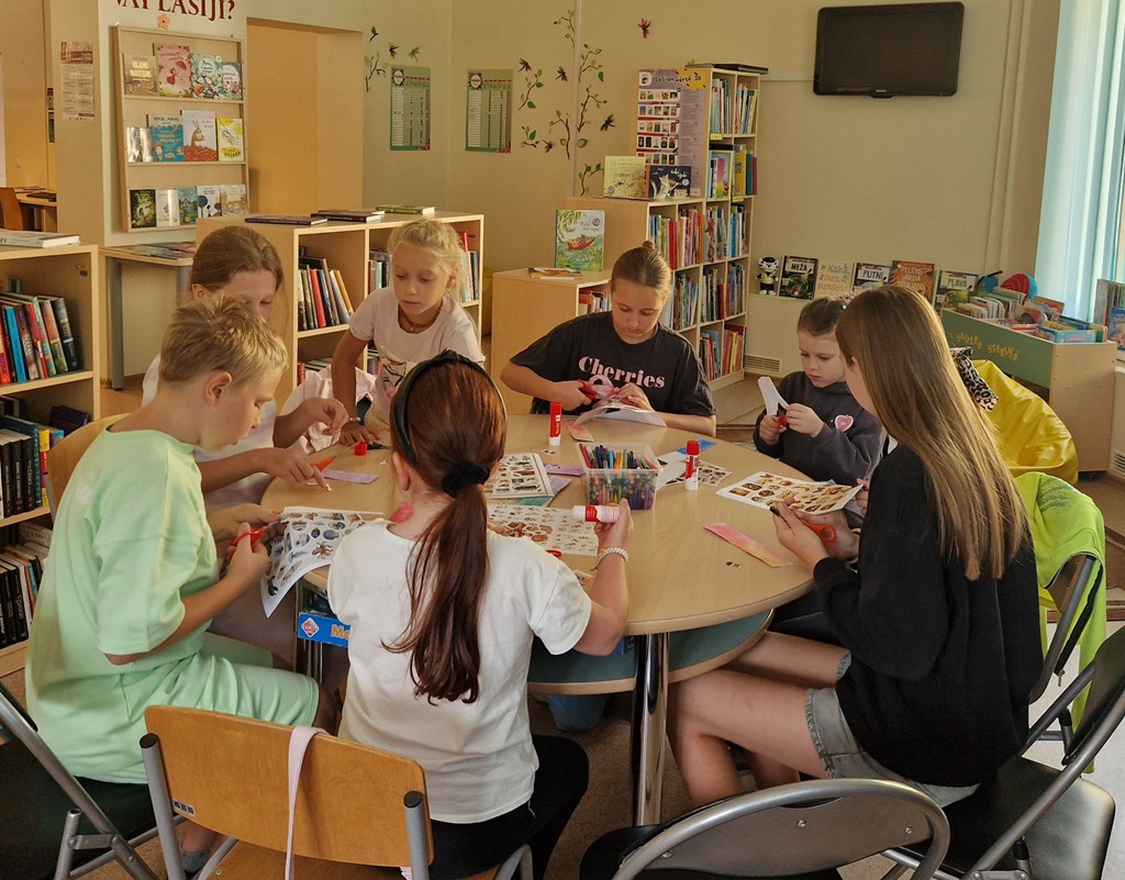 Bērni sēž ap apaļo galdu un veido papīra aplikācijas bibliotēkas radošajā darbnīcā
