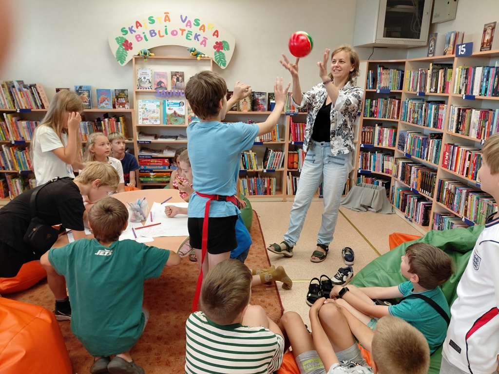 Bērni spēlē bumbas spēli ar vadītāju bibliotēkas pasākumā "Skaistā vasara"
