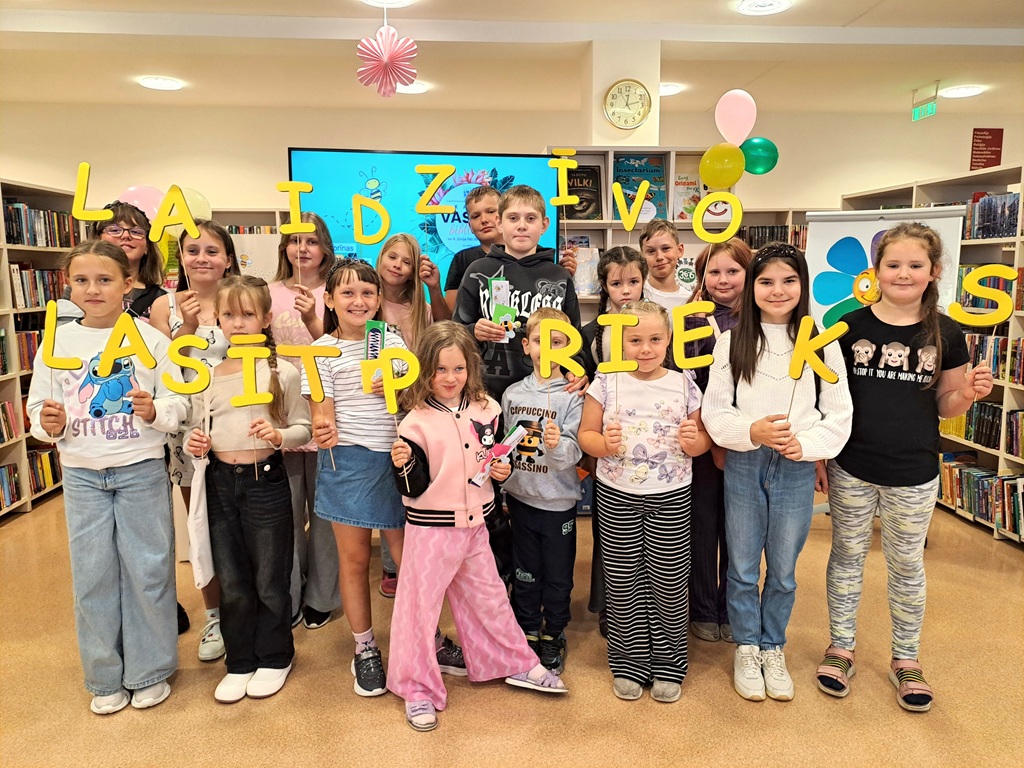 Bērni bibliotēkā tur dzeltenos burtus, veidojot uzrakstu "Lai dzīvo lasītprieks"
