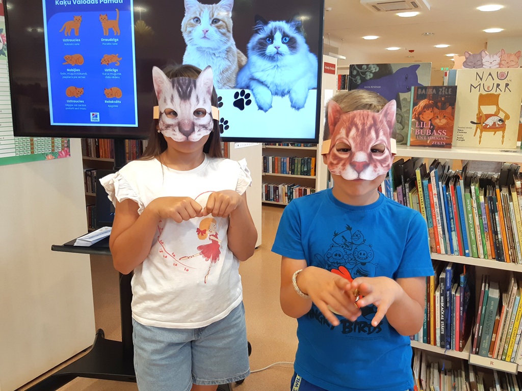 Divi bērni ar kaķu maskām pozē pie ekrāna bibliotēkas izglītojošajā nodarbībā par dzīvniekiem