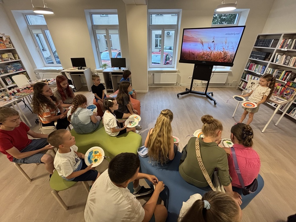 Bērni sēž puslokā un prezentē savus apgleznotos šķīvjus bibliotēkā