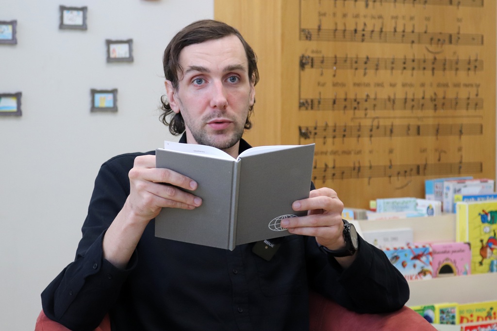 Vīrietis melnā kreklā uzmanīgi pārlūko pelēku grāmatu pasākumā bibliotēkā