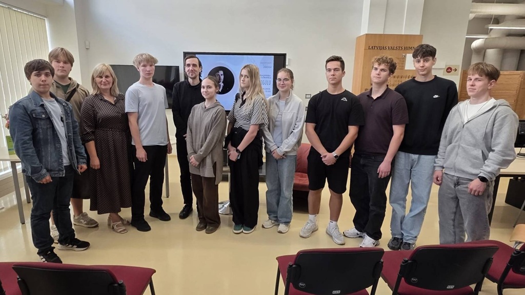 Grupas foto ar pasākuma dalībniekiem un runātāju bibliotēkas zālē pie ekrāna