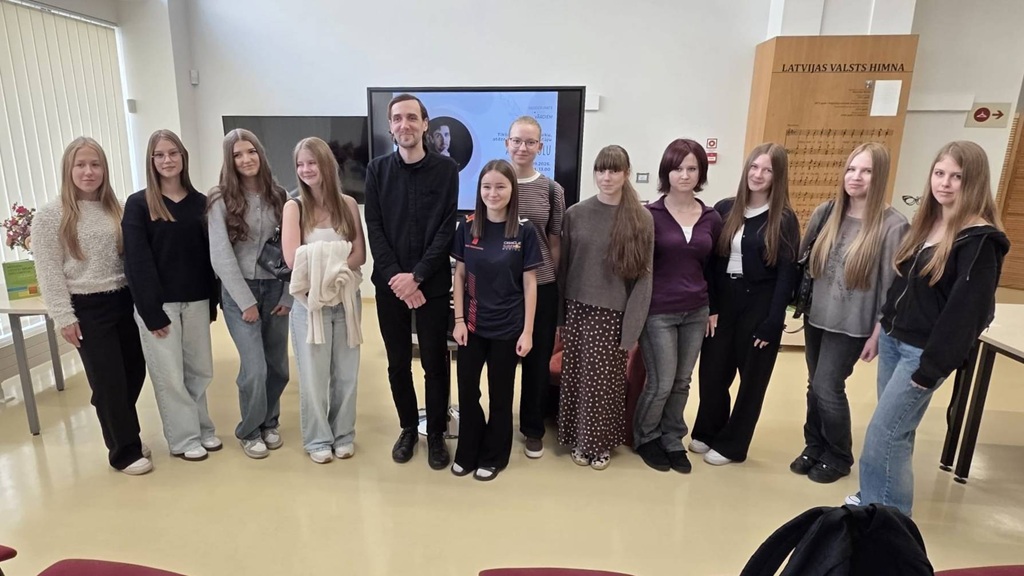 Kopīgs grupas foto ar pasākuma dalībniekiem un runātāju bibliotēkas telpā pie prezentāciju ekrāna