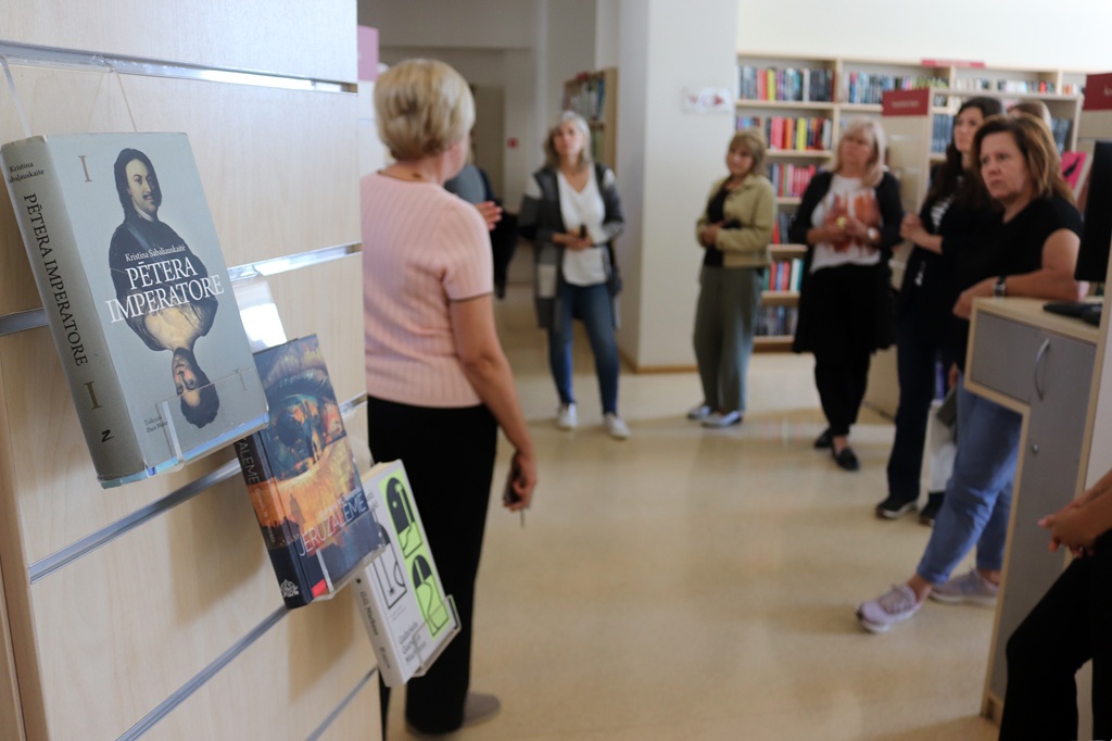 Ekskursijas dalībnieki klausās stāstījumā bibliotēkā, priekšplānā izcelta Kristinas Sabaliauskaites grāmata "Pētera imperatore"