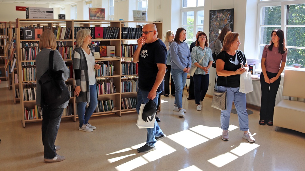 Apmeklētāju grupa sarunājas un aplūko grāmatu plauktus saulainā bibliotēkas telpā