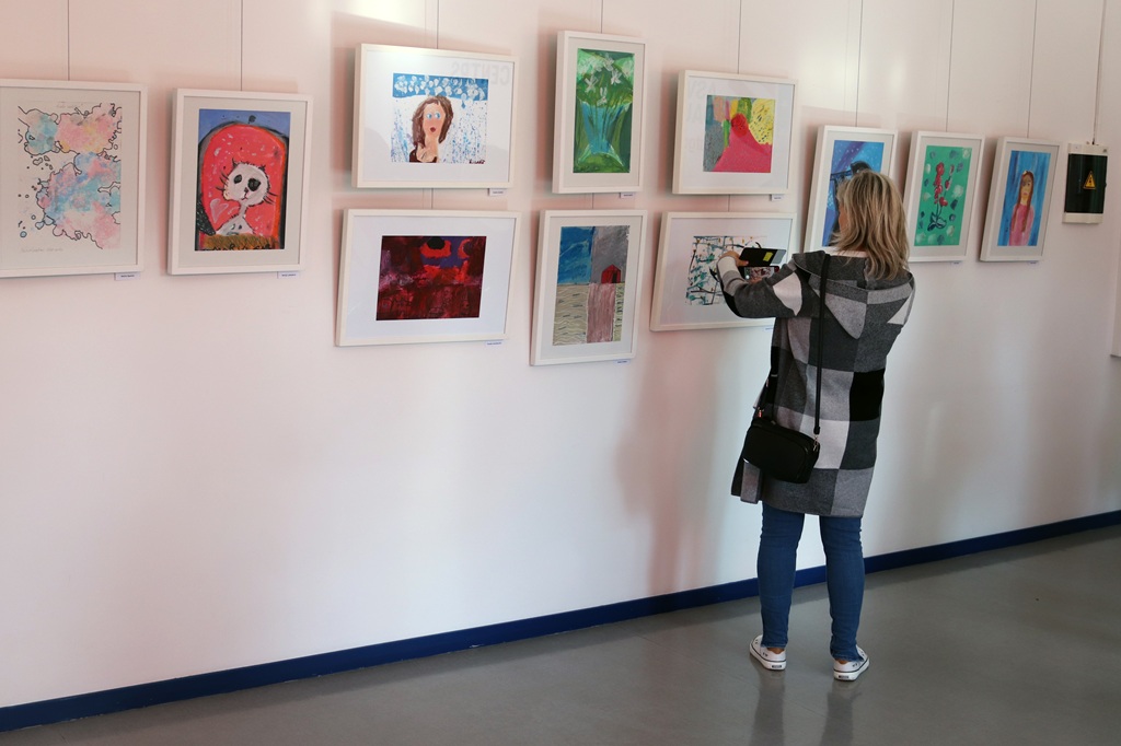Sieviete ar viedtālruni fotografē ierāmētus krāsainus zīmējumus mākslas izstādē bibliotēkā