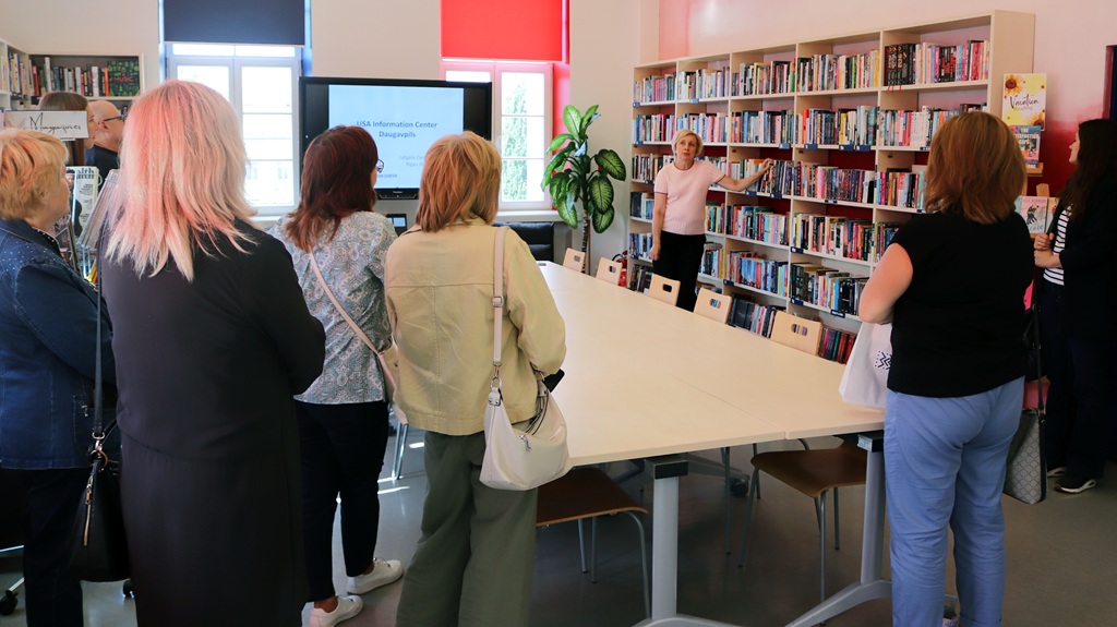 Bibliotēkas darbiniece rāda grāmatu plauktus apmeklētāju grupai ASV Informācijas centrā