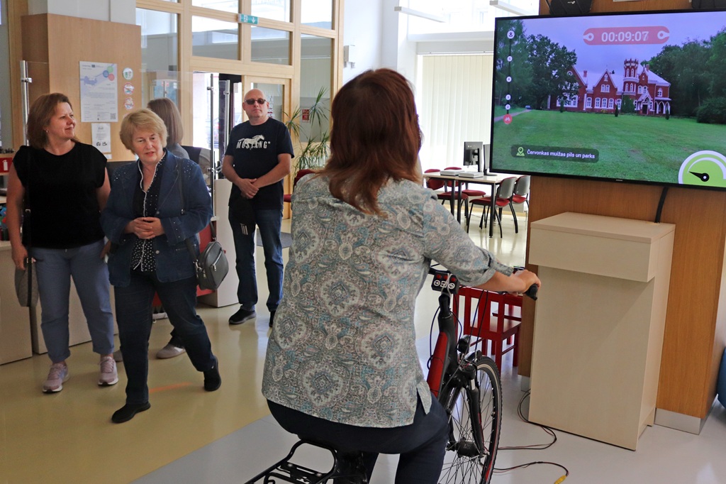 Bibliotēkas apmeklētāja brauc ar interaktīvo velosipēdu, ekrānā vērojot Červonkas muižas pili un parku.