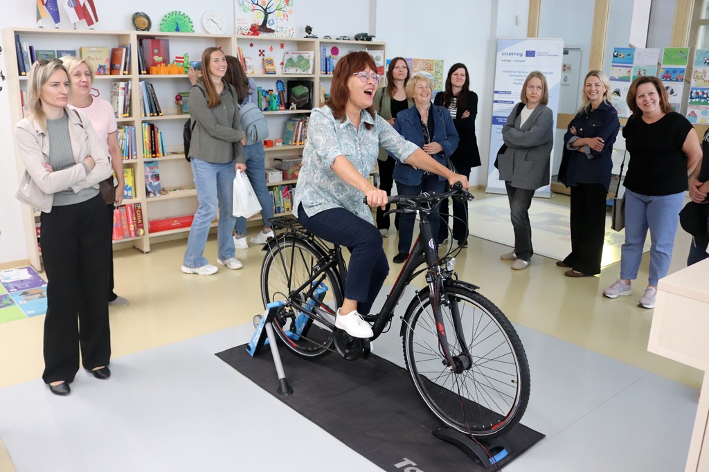 Smaidīga sieviete izmēģina interaktīvo velosipēdu grāmatu un spēļu plauktu fonā.
