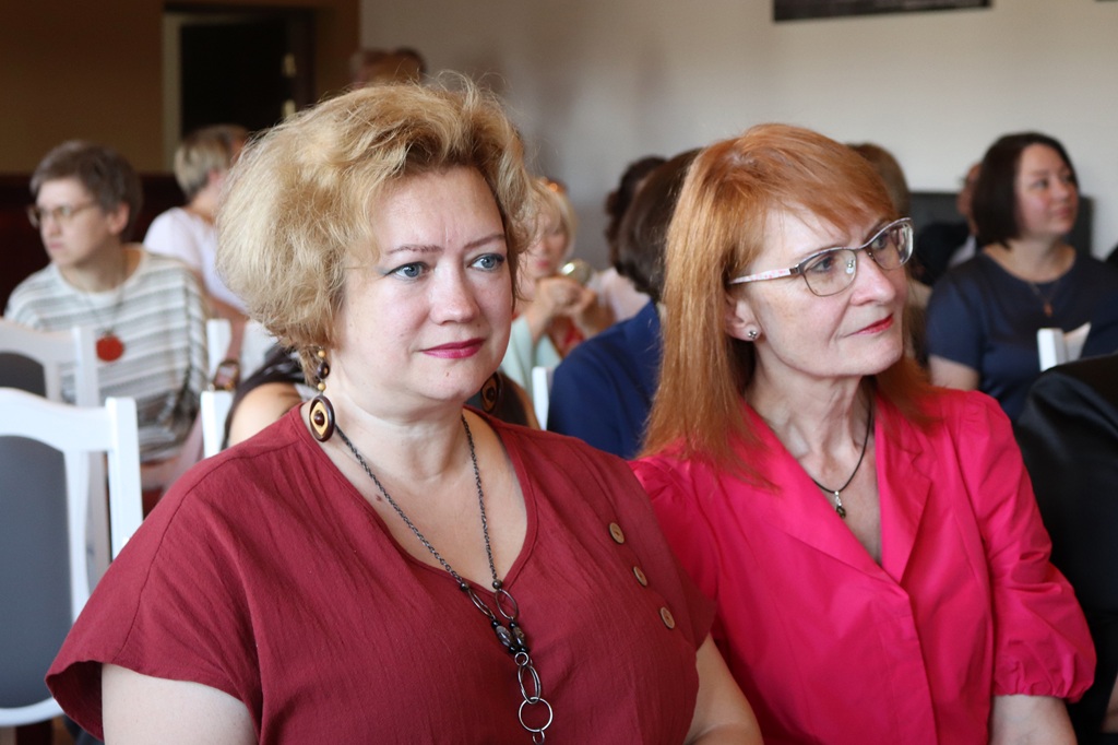 Divas sievietes sarkanā un koši rozā apģērbā uzmanīgi vēro pasākumu auditorijā
