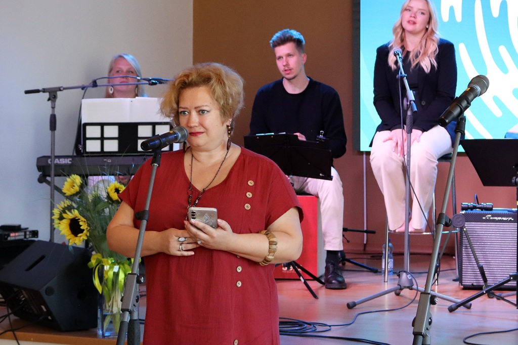 Sieviete sarkanā tērpā ar tālruni rokā runā mikrofonā, fonā mūziķu grupa