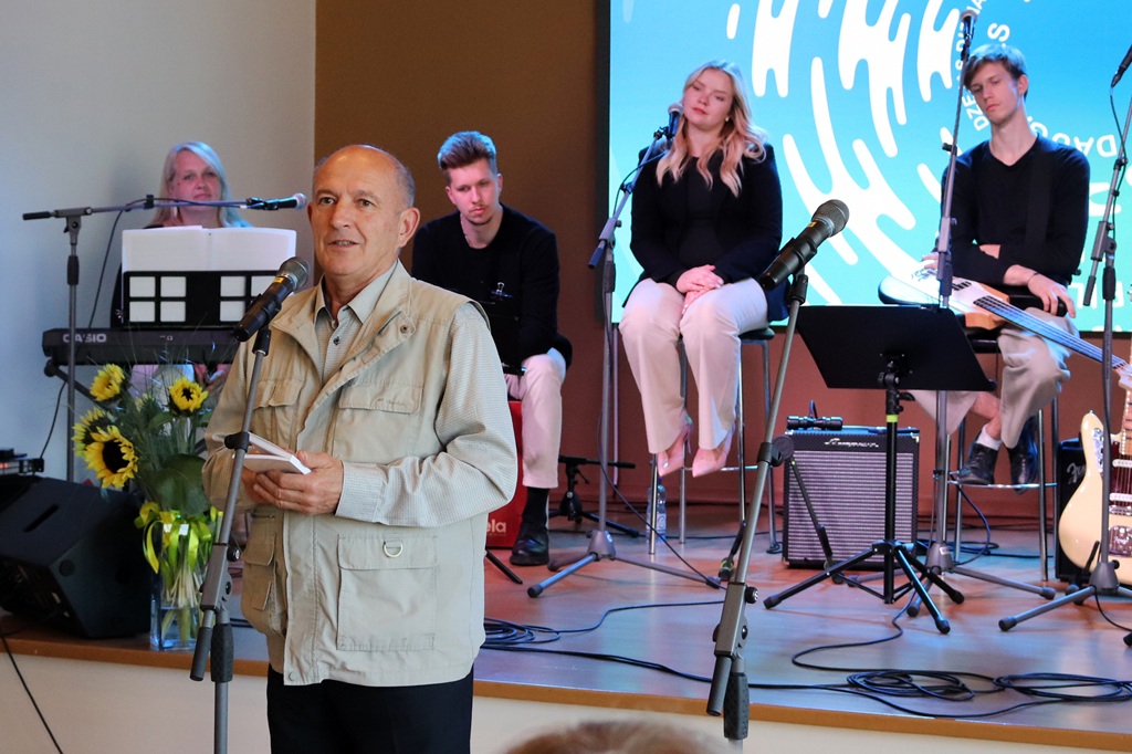 Vīrietis gaišā žaketē ar pierakstu kladi rokās uzstājas pie mikrofona, fonā mūziķu grupa