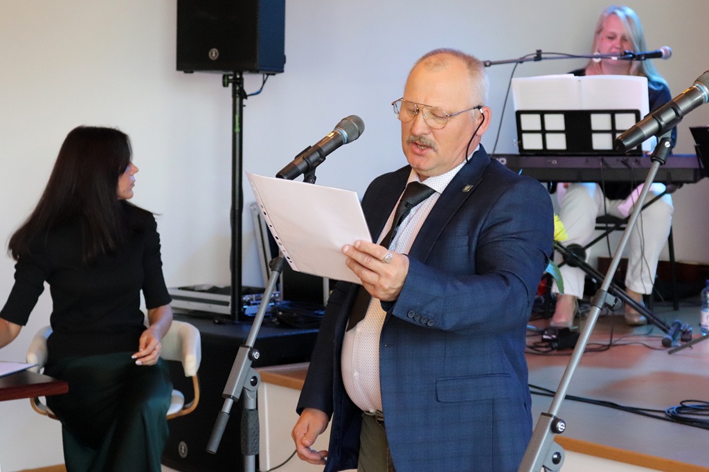 Vīrietis uzvalkā ar dokumentu mapi rokās lasa runu pie mikrofona pasākumā