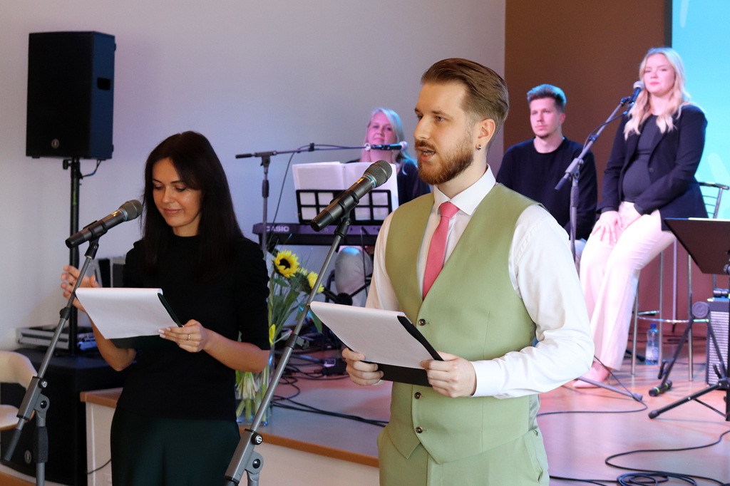 Divi pasākuma vadītāji ar lapām un mikrofoniem rokās vada pasākumu, fonā mūziķu grupa