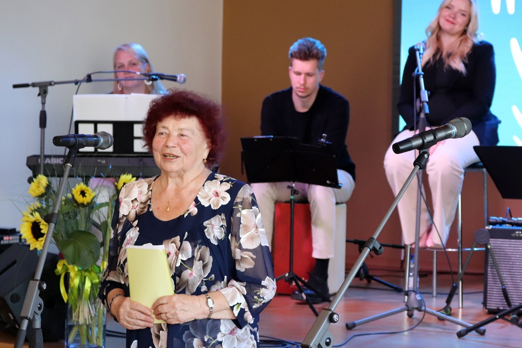 Seniora gadagājuma sieviete ziedainā blūzē ar lapu rokās smaidot uzstājas pasākumā