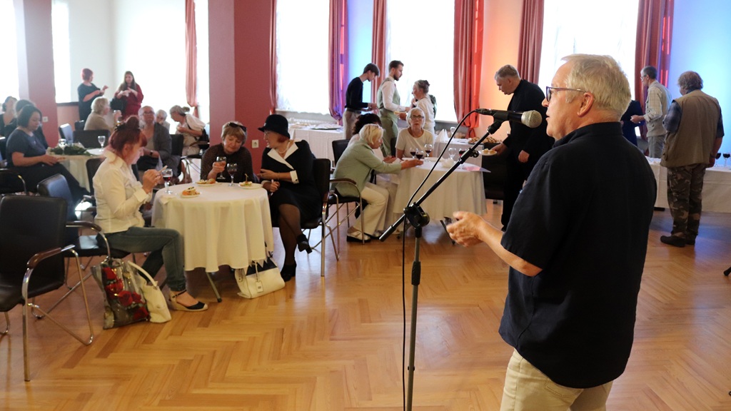 Vīrietis tumšā kreklā runā mikrofonā, amfiteātra tipa zālē pie galdiņiem sēžot pasākuma apmeklētājiem