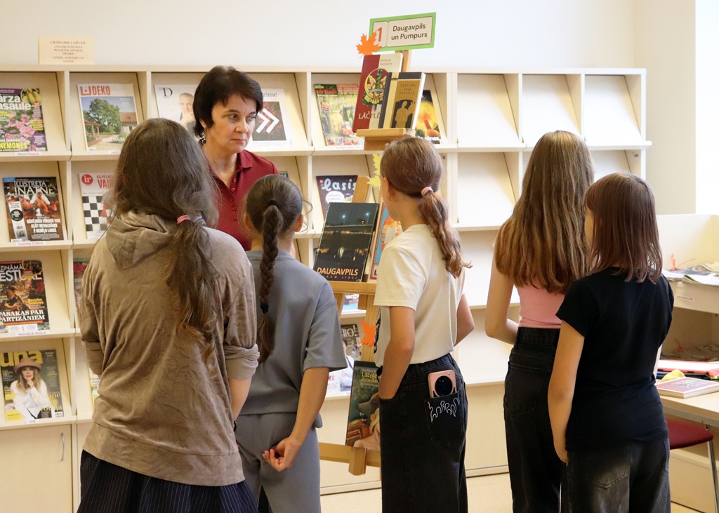 Bibliotēkas darbiniece vada dzejas stacijas uzdevumu jauniešu grupai pie molberta un žurnālu plauktiem