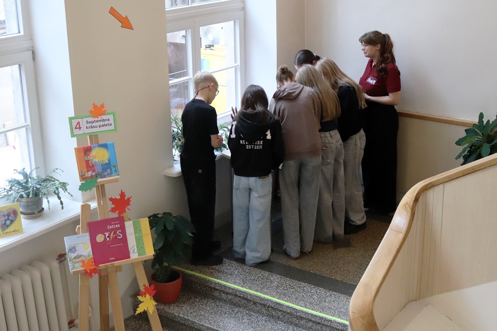 Skolēnu grupa pilda uzdevumu pie palodzes 4. stacijā "Septembra krāsu palete" bibliotēkas kāpņu laukumā