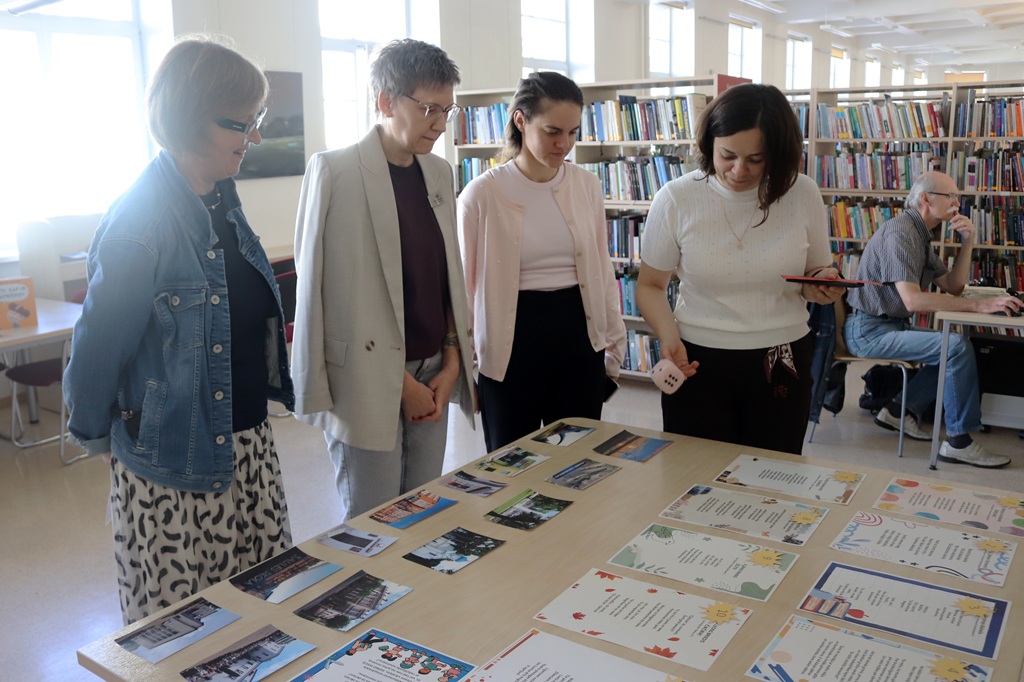 Četras sievietes pēta uz galda izkārtotās fotogrāfijas un uzdevumu lapas bibliotēkā
