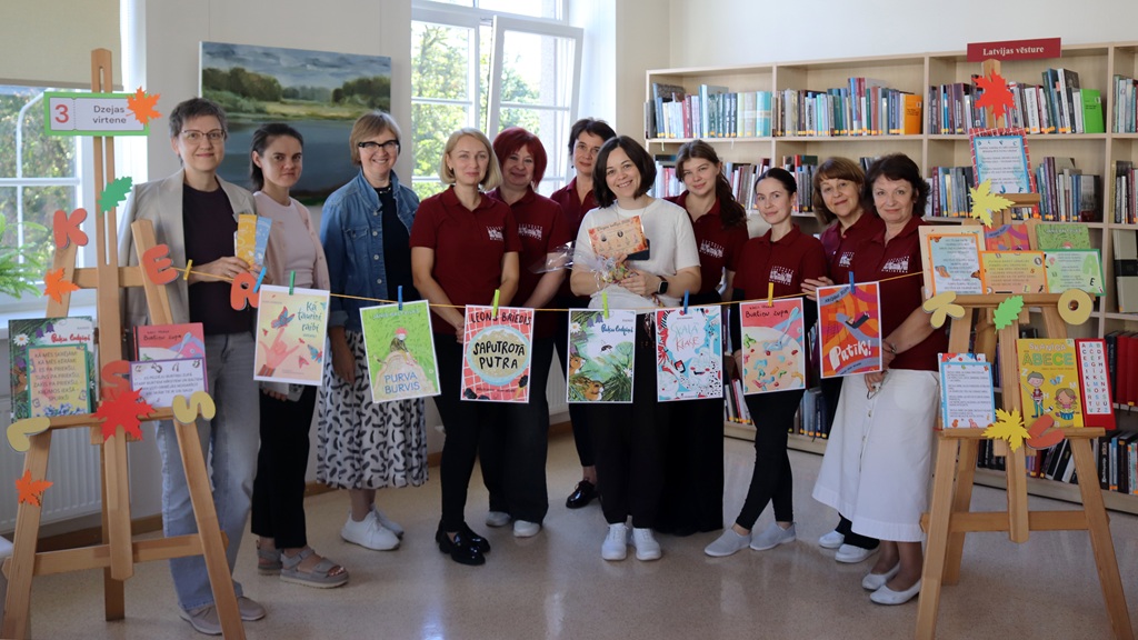 Bibliotēkas darbinieces un pasākuma organizatores kopbildē aiz auklas ar piestiprinātiem grāmatu vākiem