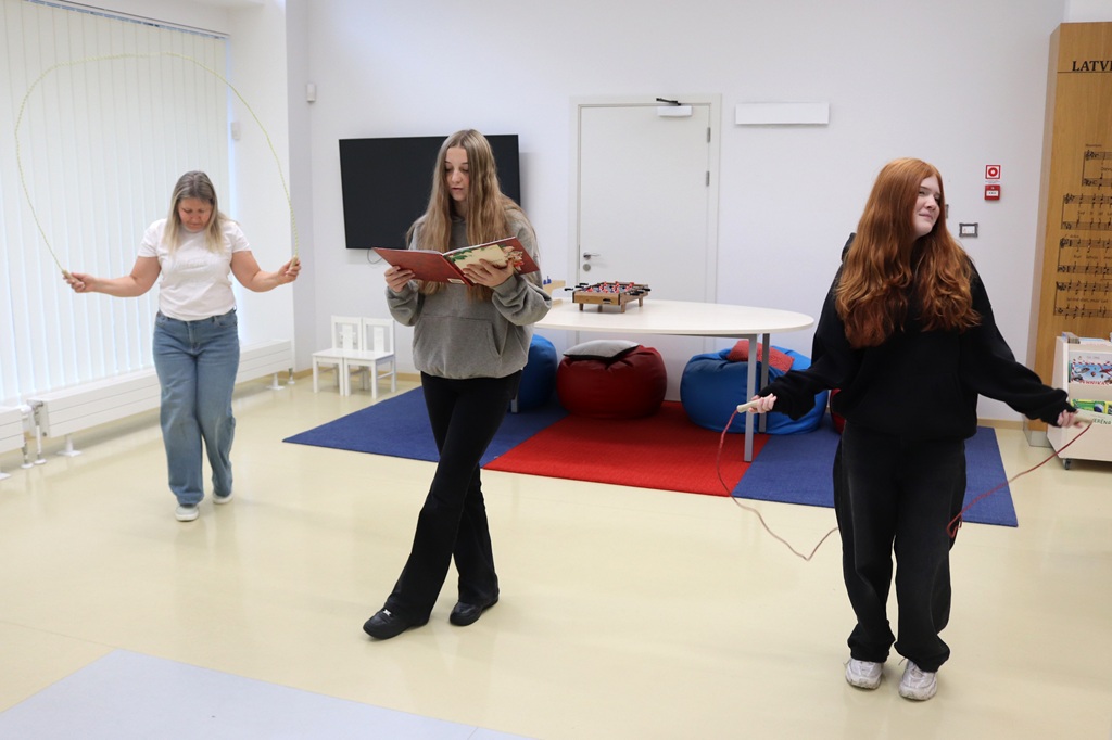 Trīs dalībnieces bibliotēkas telpā apvieno lasīšanu un lekšanu ar lecamauklu dzejas stacijas uzdevuma laikā