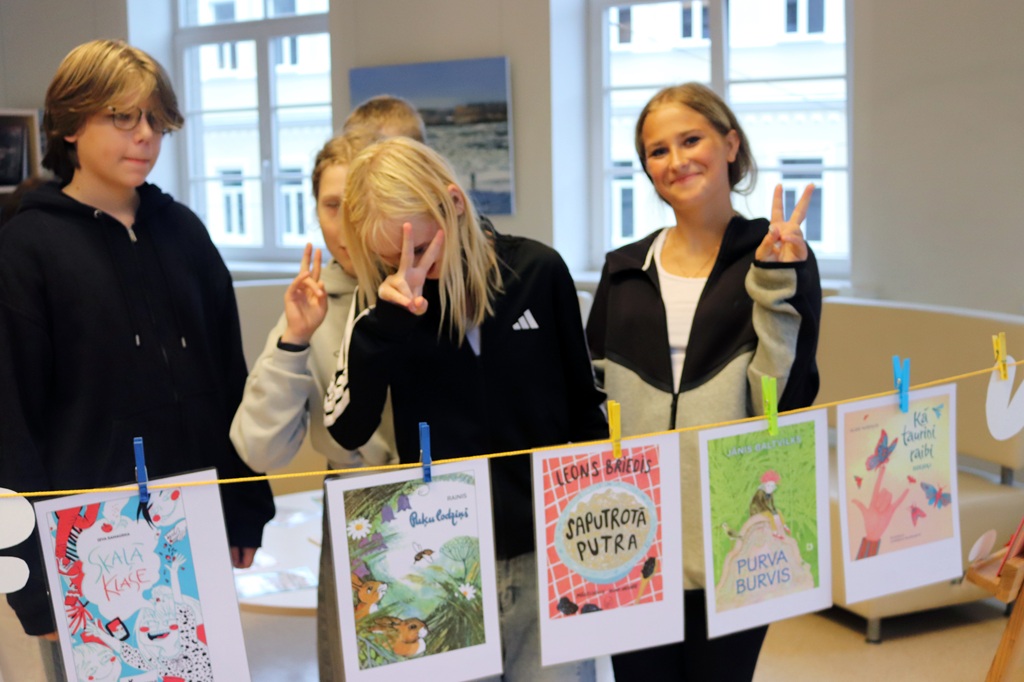 Jauniešu grupa pozē aiz veļas auklas ar piestiprinātiem grāmatu vāku attēliem