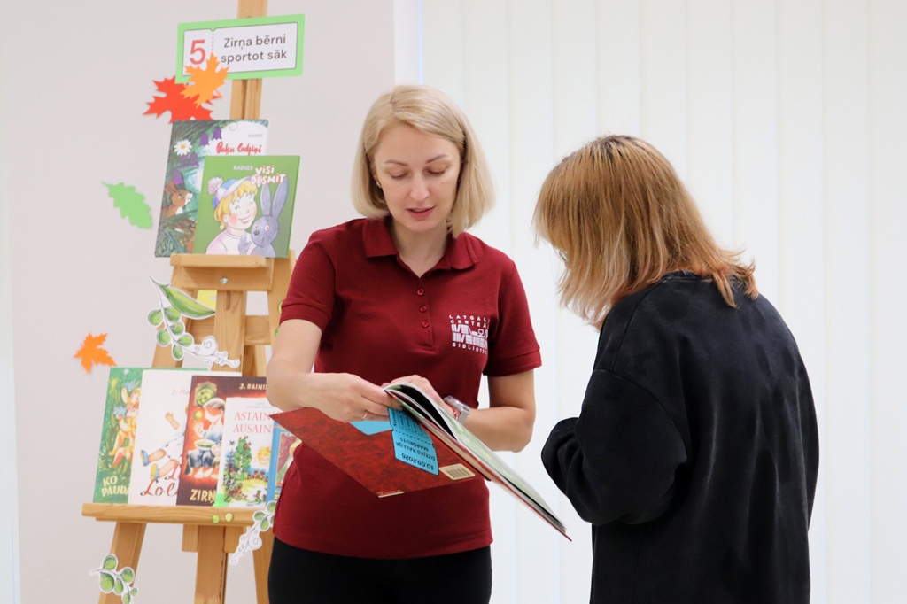 Bibliotēkas darbiniece bordo krekliņā uzrāda grāmatu un uzdevuma kartīti pasākuma dalībniecei