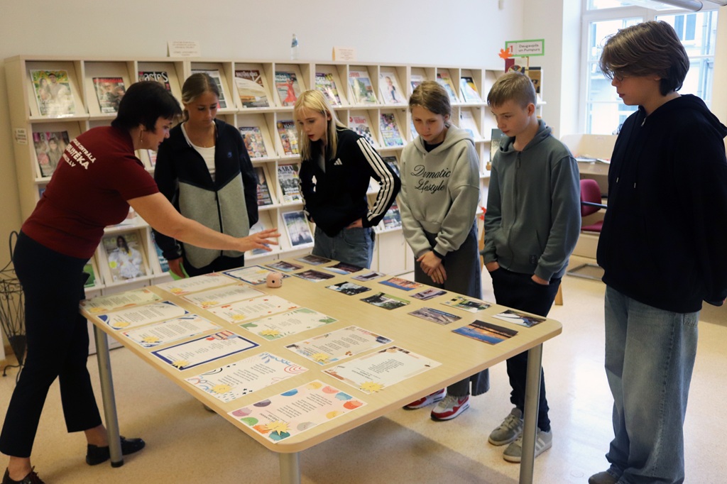 Bibliotēkas darbiniece iepazīstina jauniešu grupu ar uzdevumu lapām un fotogrāfijām uz galda