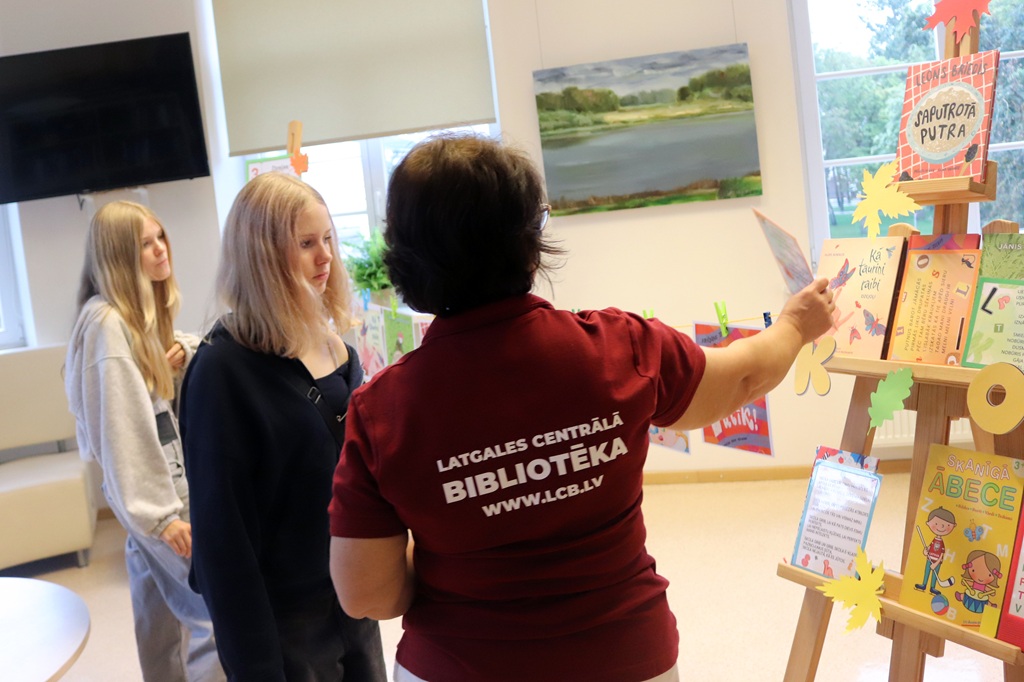 Bibliotēkas darbiniece uzstādījumā uz molberta rāda grāmatas divām jaunietēm