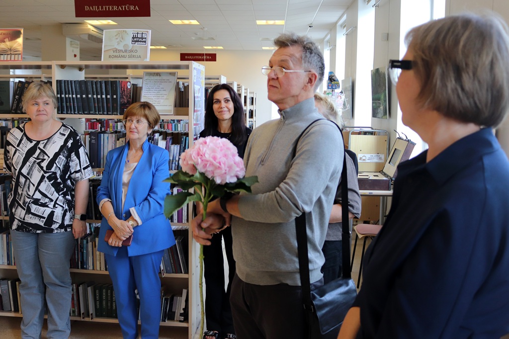 Vīrietis ar rozā hortenzijas ziedu rokās klausās uzrunu kopā ar citiem apmeklētājiem bibliotēkas dailliteratūras nodaļā