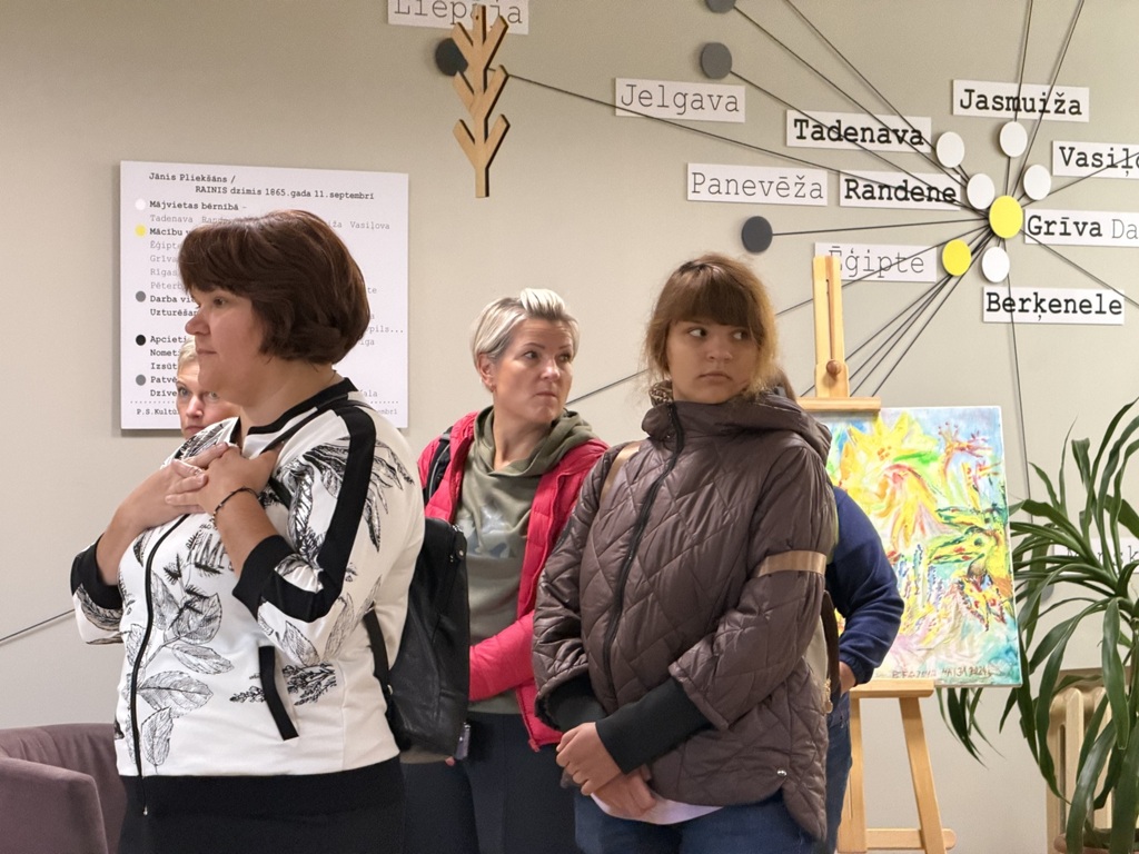 Apmeklētāji vēro izstādi pie sienas ar ģeogrāfisko vietu nosaukumiem un biogrāfisku informāciju