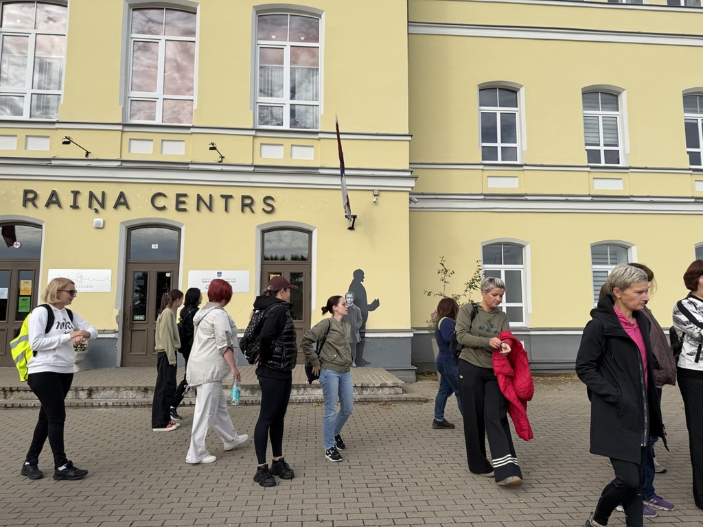 Pasākuma dalībnieki pulcējas ārpusē pie dzeltenas ēkas ar uzrakstu "Raiņa centrs"