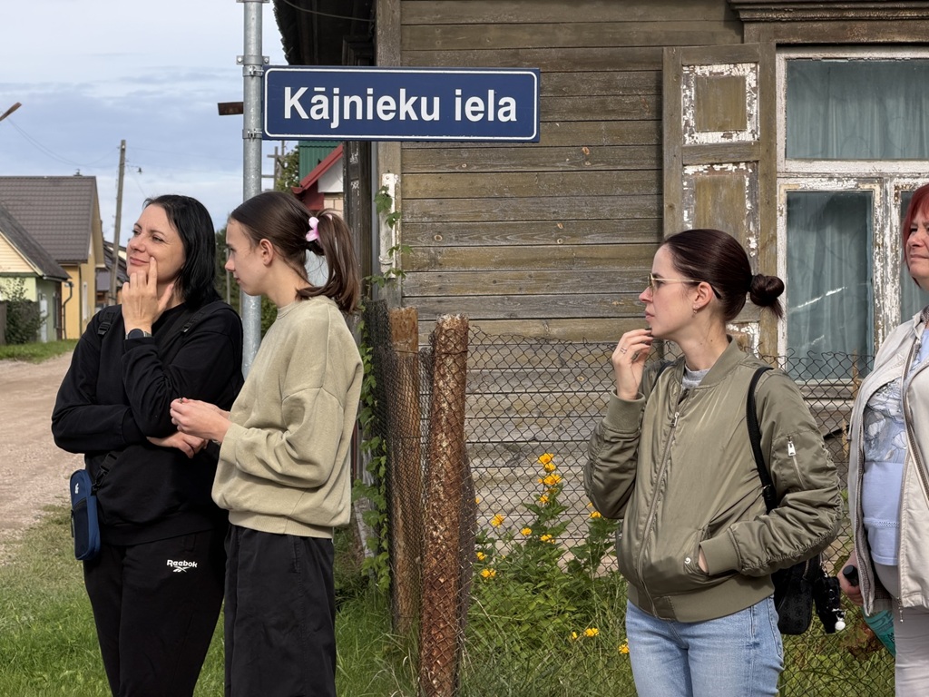 Sievietes stāv zem ielas nosaukuma norādes "Kājnieku iela" pie koka ēkas