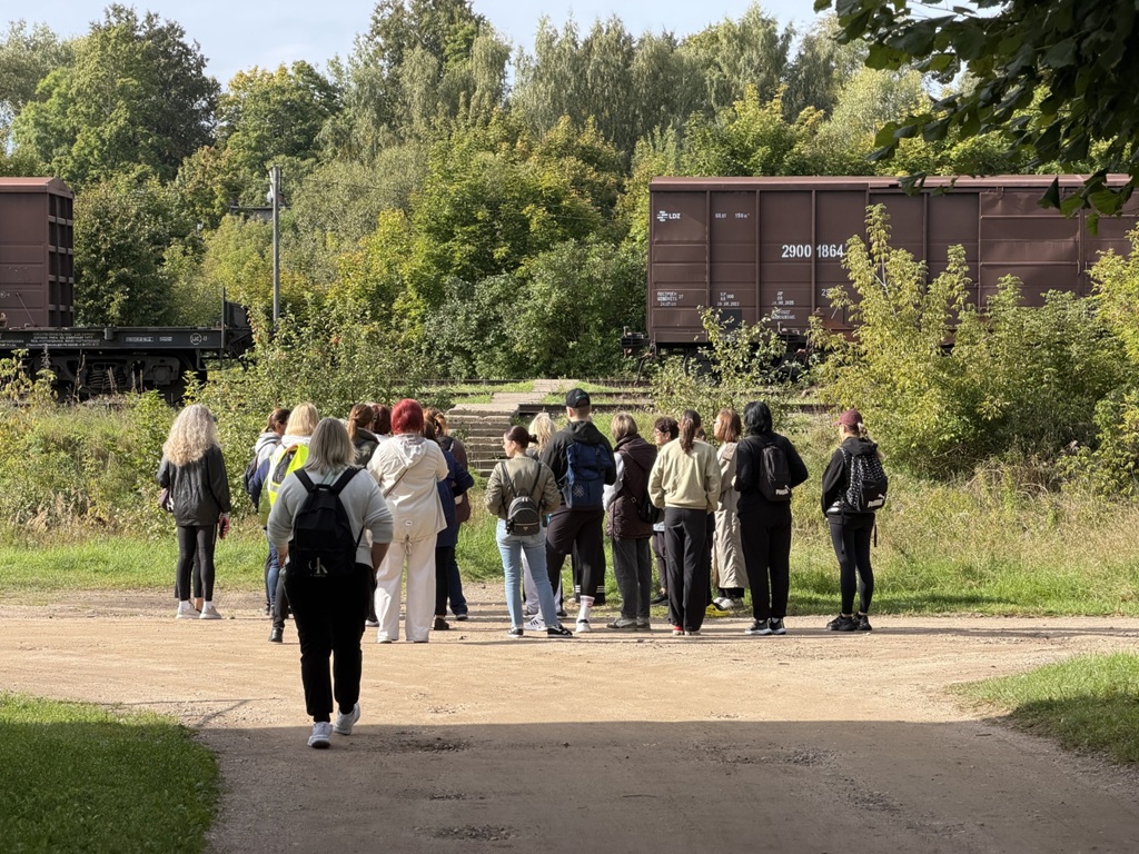 Cilvēku grupa no mugurpuses stāv uz ceļa pretī dzelzceļa vagoniem dabā