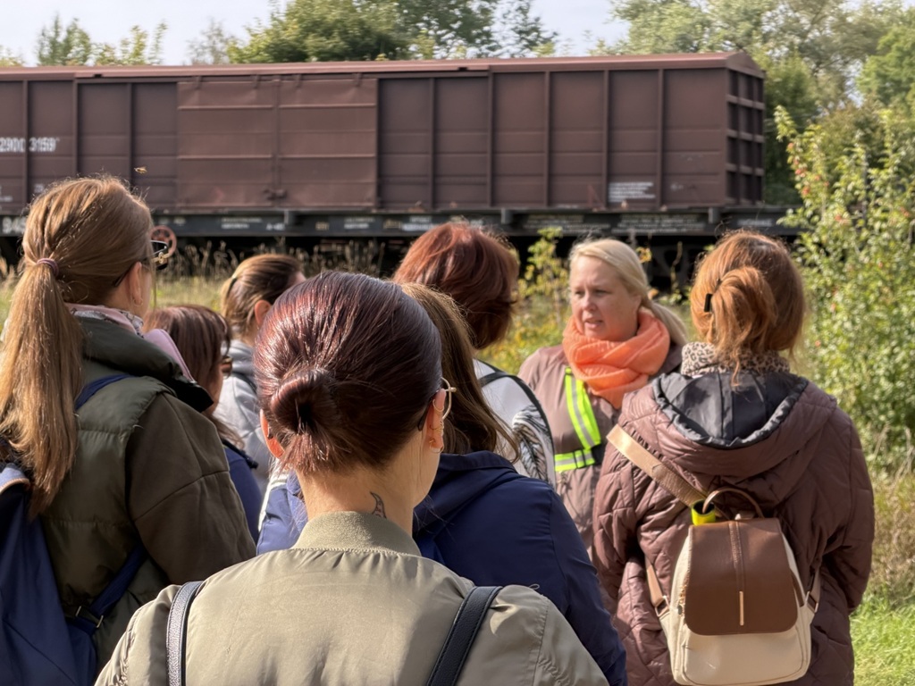 Sieviešu grupa no mugurpuses klausās gidi dabā pie dzelzceļa vagona