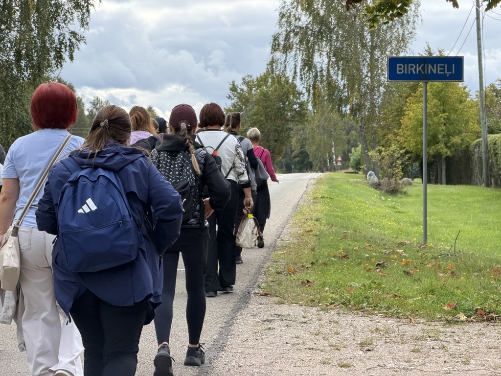 Cilvēku grupa iet gar Birķineļu ciema zīmi lauku ceļā