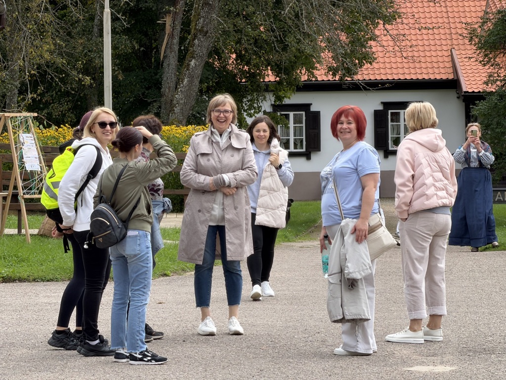 Cilvēku grupa pie tradicionālās lauku mājas ar sarkano jumtu