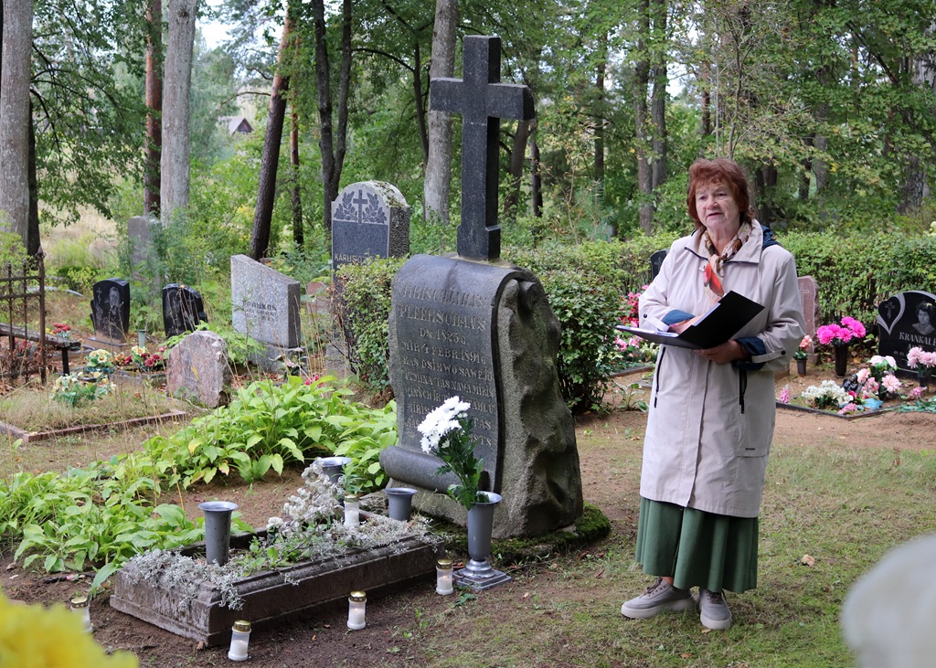 Sieviete gaišā mētelī lasa no mapes pie vēsturiska akmens pieminekļa ar krustu kapsētā