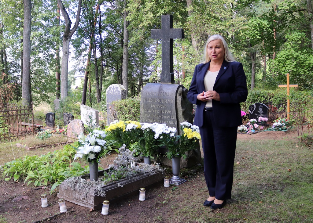 Sieviete tumšā kostīmā uzstājas pie ziediem un svecēm rotātas kapa vietas ar akmens krustu