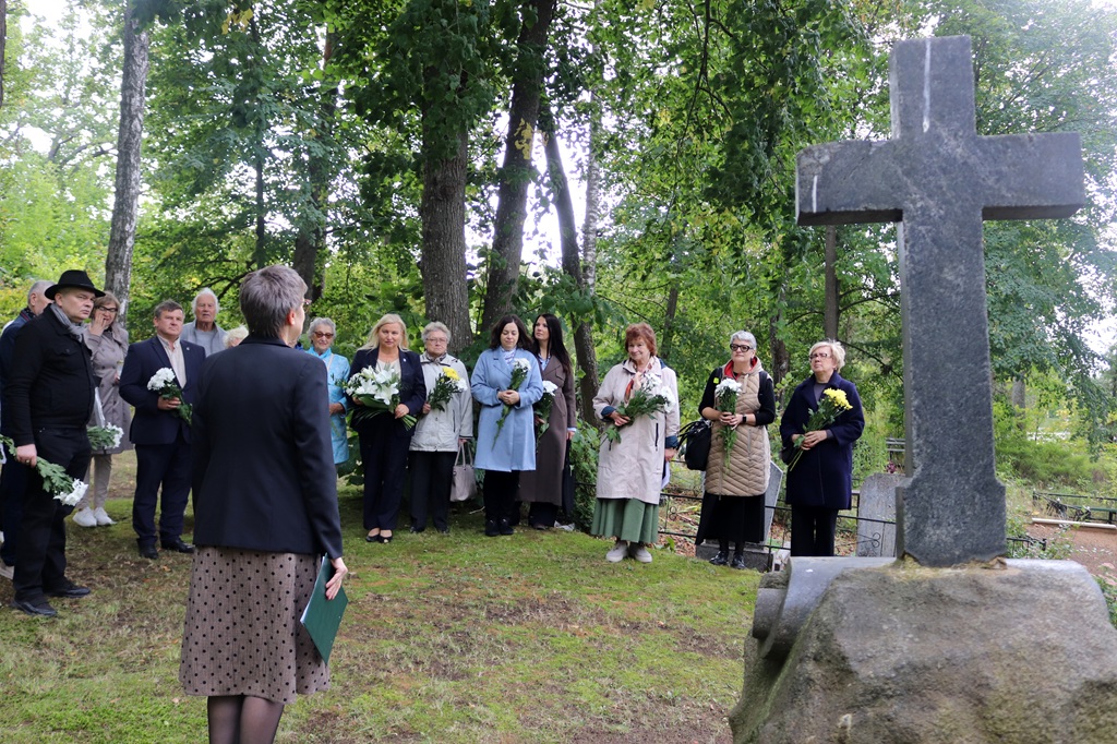Sieviete no mugurpuses uzrunā pasākuma dalībniekus pie akmens krusta kapsētā
