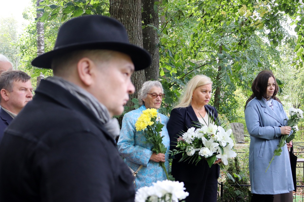 Pasākuma dalībnieces ar baltiem un dzelteniem ziedu pušķiem stāv piemiņas brīdī kapsētā