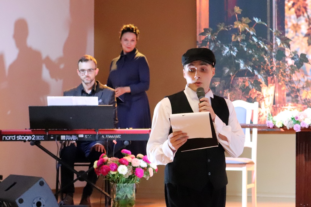 Jauns vīrietis cepurē un vestē uzstājas pie mikrofona ar mapi rokās, fonā pianistis un sieviete